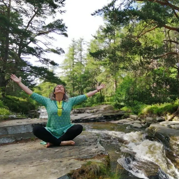 An adult lady sitting in Meditation & Yin Yoga sequence for hips pose, surrounded by their background trees & flowing stream