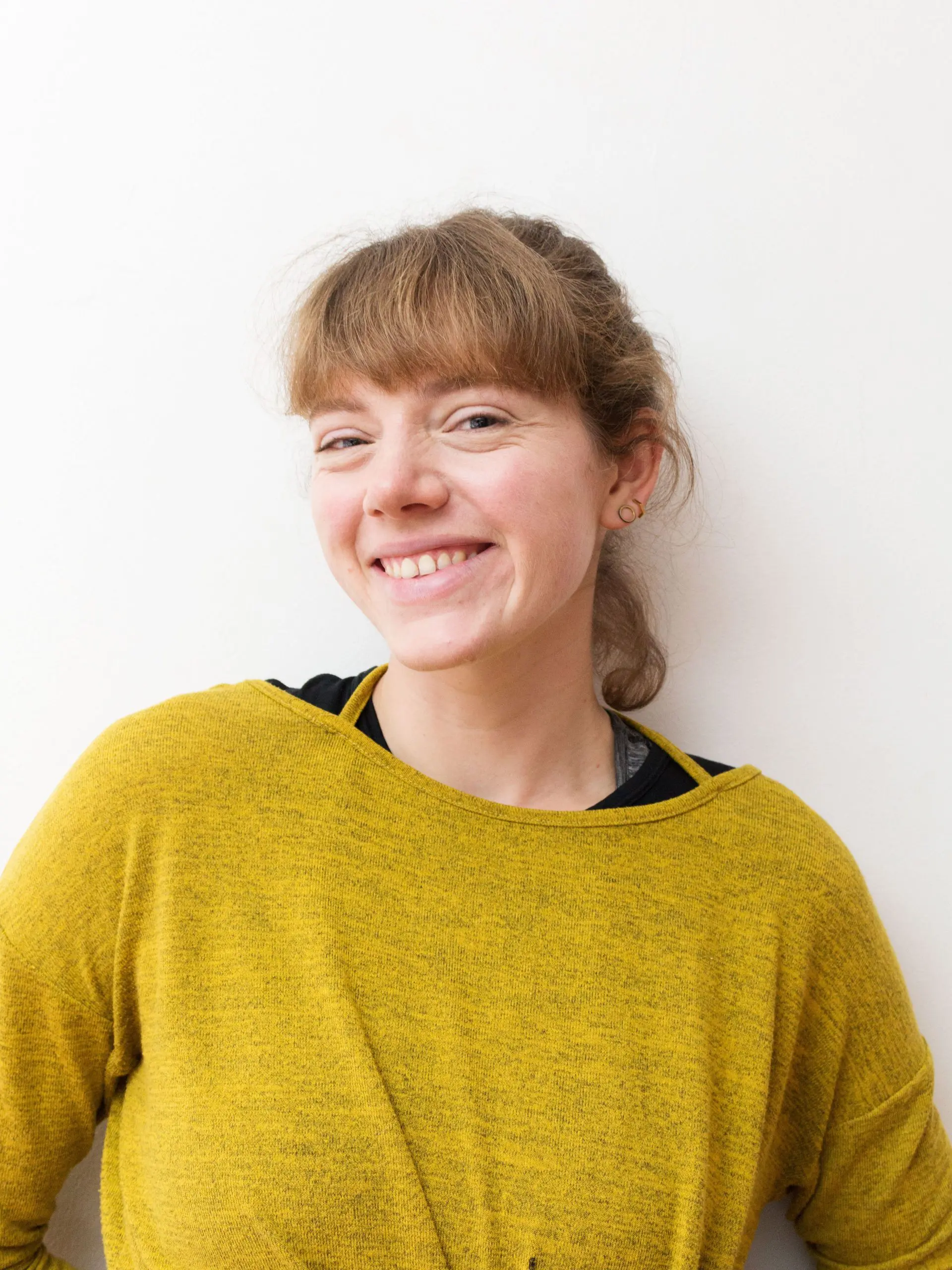 An adult lady who has brown hair and wearing a yellow shirt leans against a white wall | The Helpful Yoga Poses